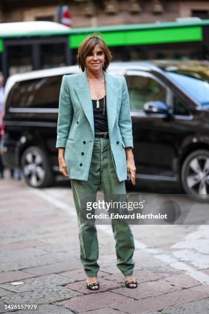Carine Roitfeld wears gold earrings, gold large chain necklaces, a pale green long wool blazer jacket, a black V-neck tank-top, a black shiny leather...