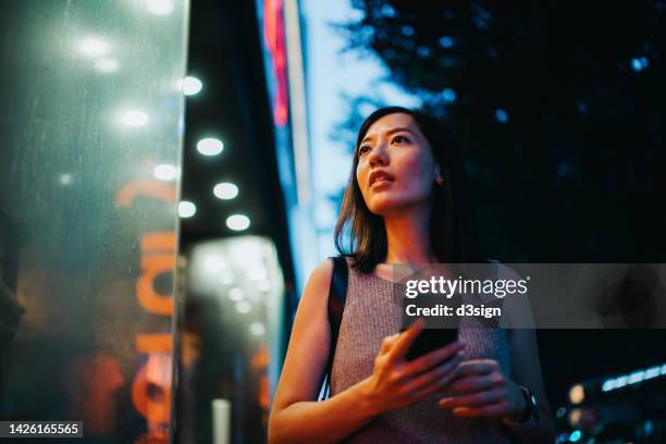 young asian woman using smartphone while looking at shop window outside a fashion boutique, walking in city in the evening, with colourful neon lighting reflected on her - angeleuchtet zahlen mensch stock-fotos und bilder