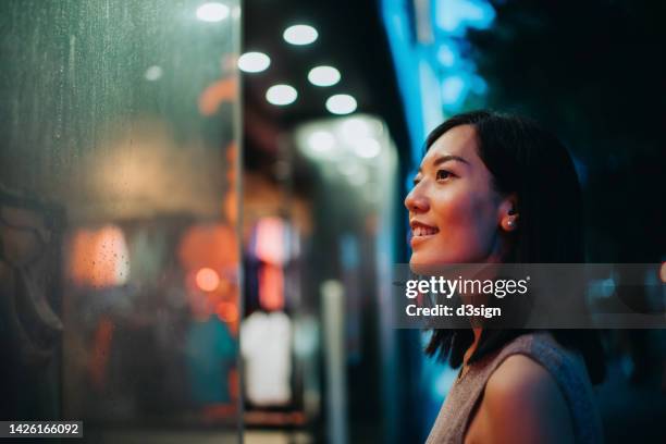 smiling young asian woman standing in front of a boutique and looking at shop window, shopping in city in the evening, with colourful neon lighting reflected on her - ショーウィンドウ ストックフォトと画像