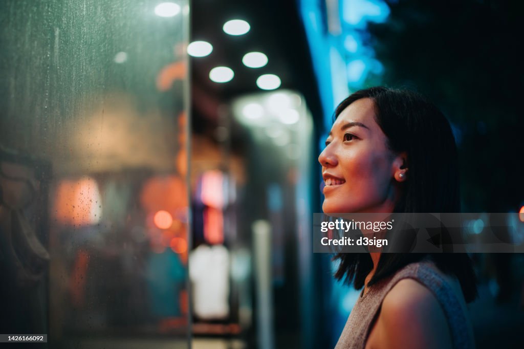 Smiling young Asian woman standing in front of a boutique and looking at shop window, shopping in city in the evening, with colourful neon lighting reflected on her