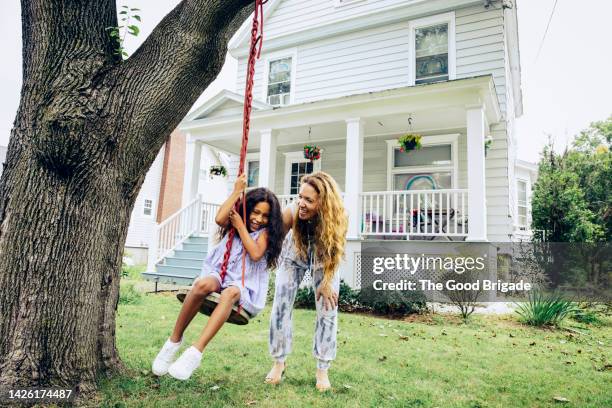 happy mother pushing daughter on swing in front yard at home - stéréotype de la classe moyenne photos et images de collection