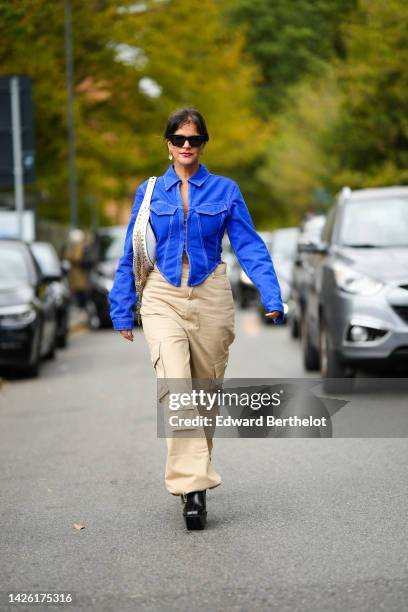 Gili Biegun wears black sunglasses, a royal blue denim asymmetric cropped / long sleeves jacket, a white shiny leather shoulder bag with nailed /...