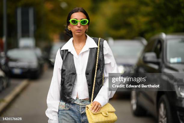 Gabriella Berdugo wears neon green sunglasses, a white shirt, a black shiny leather sleeveless buttoned gilet, a pale yellow and white tweed shoulder...