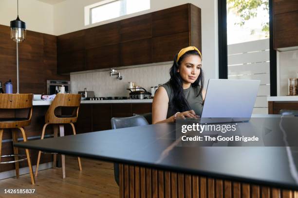 woman using laptop sitting at dining table - woman portrait kitchen laptop stock-fotos und bilder