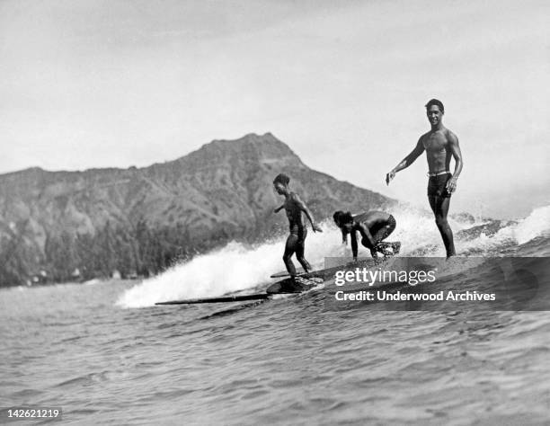 Three native surfers ride their boards with ease at Waikiki Beach, with Diamond Head in the background, Honolulu, Hawaii, 1920s.