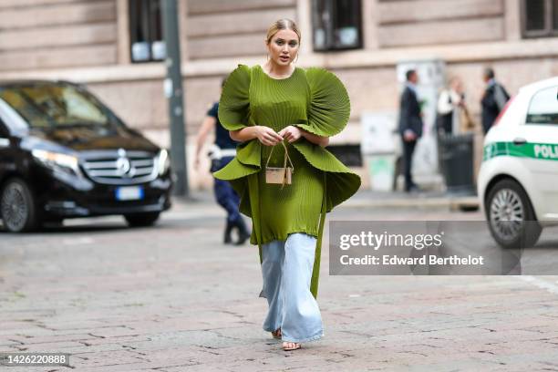 Guest wears a khaki green short dress with oversized accordion wavy short skirt, a silver metallic small handbag from Dior, gold rings, blue faded...
