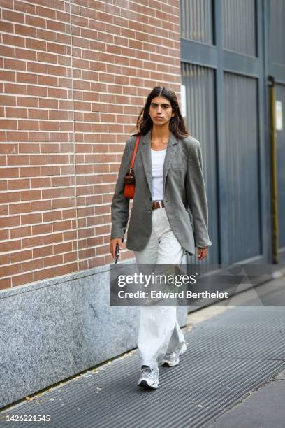 Model wears a white tank-top, a gray blazer jacket, a brown shiny leather belt, an orange shiny leather shoulder bag, white latte denim large pants,...