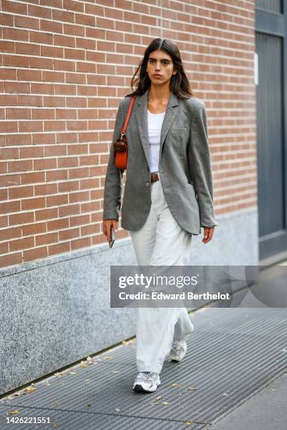 Model wears a white tank-top, a gray blazer jacket, a brown shiny leather belt, an orange shiny leather shoulder bag, white latte denim large pants,...