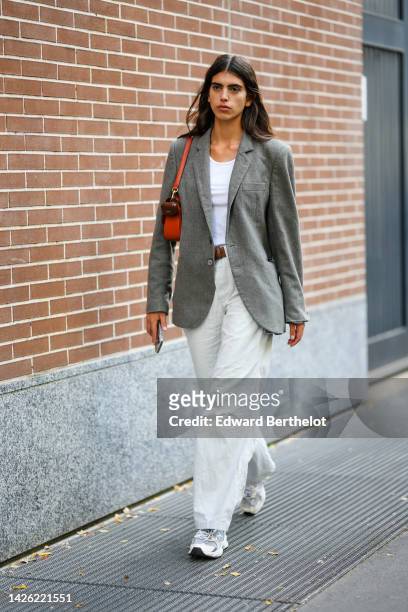 Model wears a white tank-top, a gray blazer jacket, a brown shiny leather belt, an orange shiny leather shoulder bag, white latte denim large pants,...