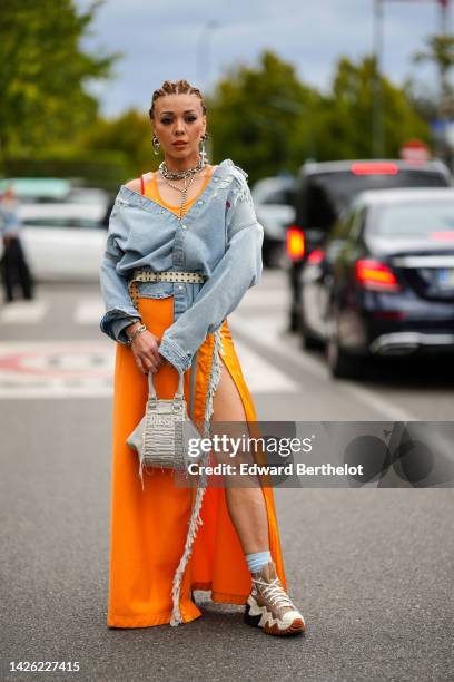 Guest wears silver large earrings, silver large necklaces, a pale gray denim ripped jacket from Diesel, an orange long dress with blue denim fringed...