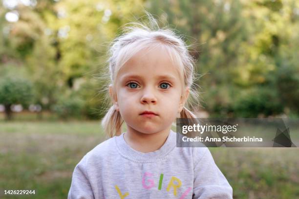 a cute blonde girl with blue eyes. the child looks at the camera. intense little girl. against the background of the park. close-up. outdoors - young face serious at camera photos et images de collection