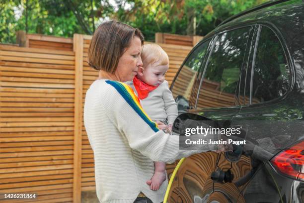 mature woman (holding her baby daughter) plugs in the electric car to charge - electric car home stock pictures, royalty-free photos & images