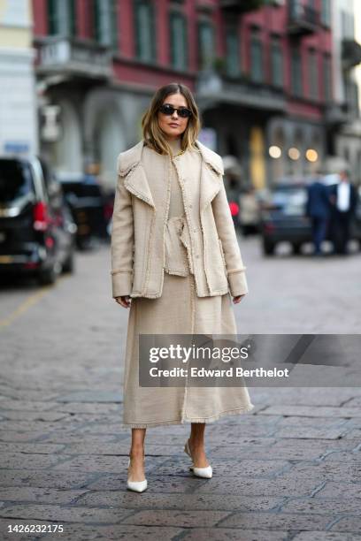 Guest wears black sunglasses, a beige turtleneck pullover, a beige tweed fringed borders double jacket, a matching beige tweed fringed borders wrap...