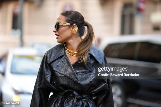 Guest wears black butterfly sunglasses, a gold large earring, a gold large chain necklace, a black shiny leather belted oversized jacket, outside...