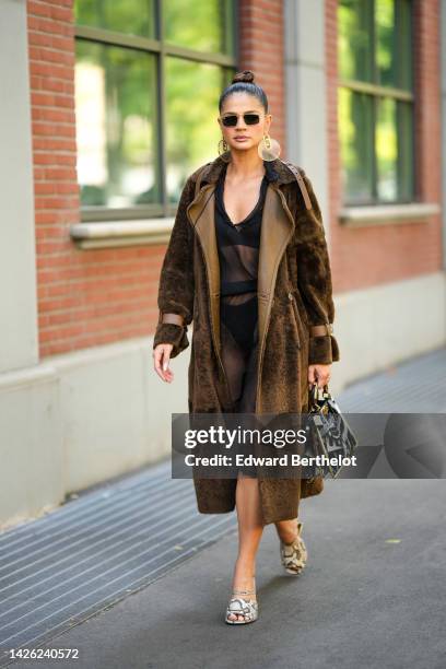 Guest wears black sunglasses, gold large circle and logo earrings from Fendi, a black silk V-neck midi dress, a brown leather and fur long coat, a...