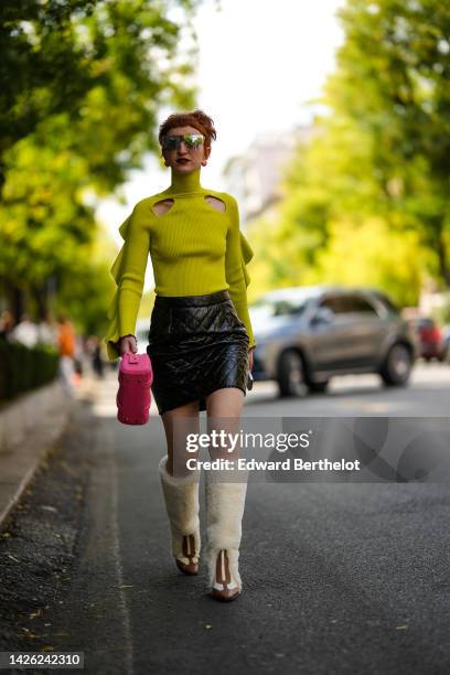 Guest wears futurist sunglasses, red and white earrings, a neon yellow turtleneck / cut-out chest / embroidered wavy long sleeves pullover, a black...