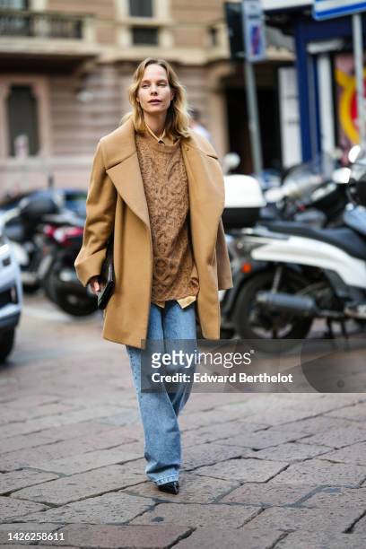 Guest wears a brown oversized felt coat, a brown braided wool oversized pullover, a brown shirt, blue faded denim large pants, a black shiny...