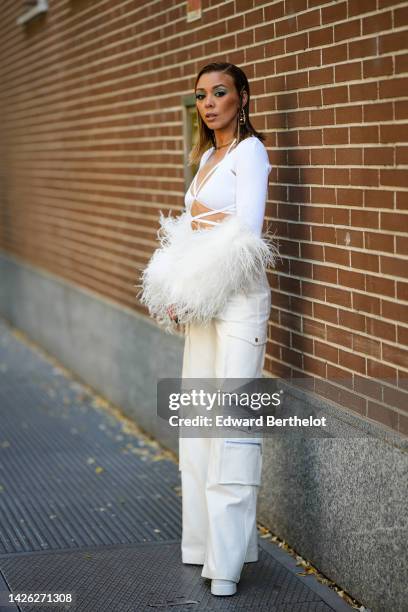 Guest wears gold large pendant logo earrings, a gold chain necklace, a white laces waits and chest / long feathers sleeves cropped top, white latte...