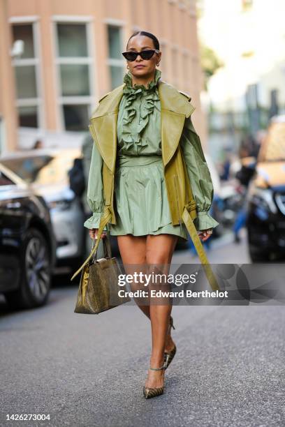 Guest wears black cat eyes sunglasses, gold and diamonds earrings, a green long puffy sleeves / ruffled belted short skirt, a khaki shiny leather...