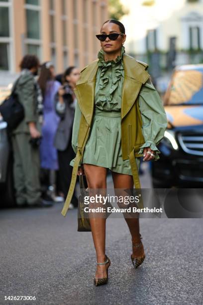 Guest wears black cat eyes sunglasses, gold and diamonds earrings, a green long puffy sleeves / ruffled belted short skirt, a khaki shiny leather...
