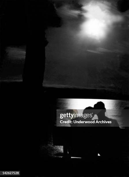 The silhouette of a couple sitting on a bench overlooking a lake in the moonlight, mid to late 1930s.