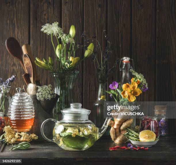 healthy herbal tea in glass teapot on dark wooden table with various ingredients: herbs, flowers, honey, lemon, and ginger. - honey lemon stock pictures, royalty-free photos & images