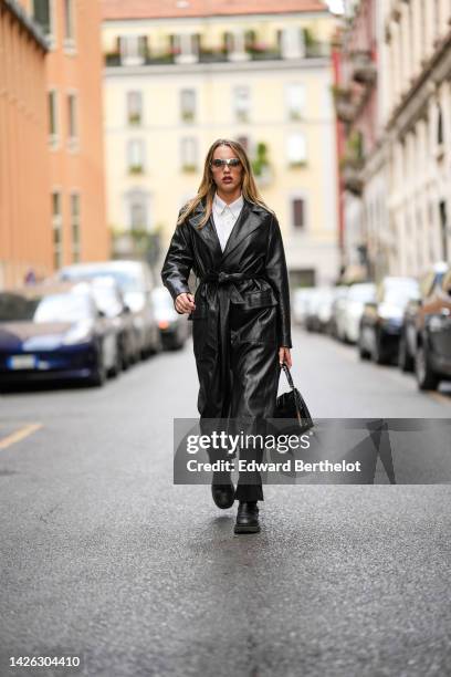 Guest wears sunglasses, a white shirt, a black shiny leather belted long trench coat, black pants, a black shiny leather handbag, black shiny leather...