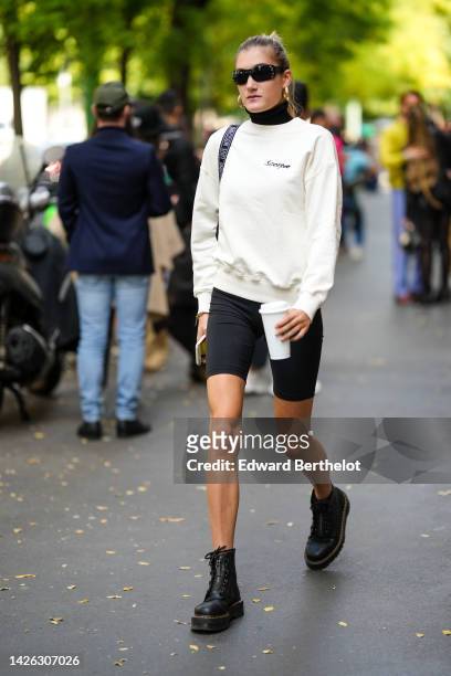 Guest wears black sunglasses, a black shoulder bag from Moschino, a black turtleneck pullover, a white latte sweater from Scorpio, black cyclist...
