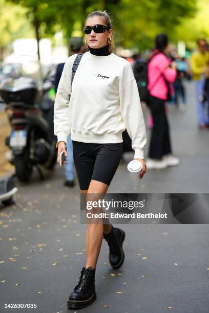 Guest wears black sunglasses, a black shoulder bag from Moschino, a black turtleneck pullover, a white latte sweater from Scorpio, black cyclist...