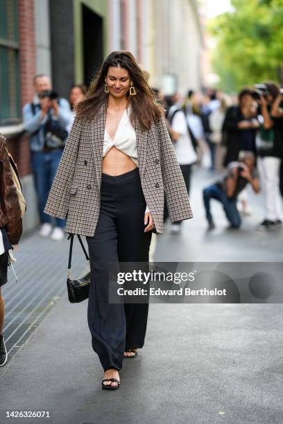 Guest wears gold large earrings, a white knot V-neck / cropped top, a brown and beige checkered print pattern blazer jacket, black large pants, a...