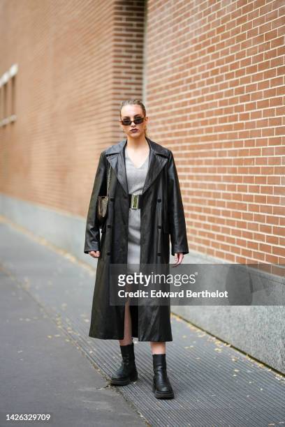 Guest wears black sunglasses, a black shiny leather long coat, a pale gray V-neck checkered print pattern short dress, a black shiny leather belt, a...