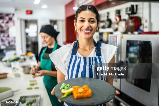portrait of a young woman showing a dish at the kitchen - happy chef stock pictures, royalty-free photos & images