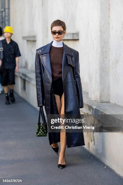 Melanie Kieback wears black oversized leather coat, white turtleneck, brown jumper, shorts, green black bag, heels outside Prada during the Milan...