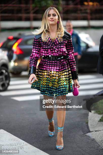 Valentina Ferragni wears pink earrings, a black with neon pink / purple / yellow / green print pattern belted coat, a neon pink shiny leather handbag...