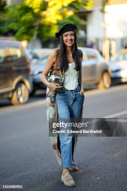 Yoyo Cao wears a black and felt with leather details beret hat, a white latte shoulder-off corset, blue faded denim straight pants, a brown with pale...