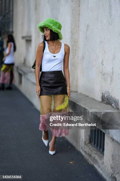 Molly Chiang wears a green fluffy bob hat, a white tank-top from Prada, a multi materials black shiny leather / yellow tulle / pink tulle long skirt...