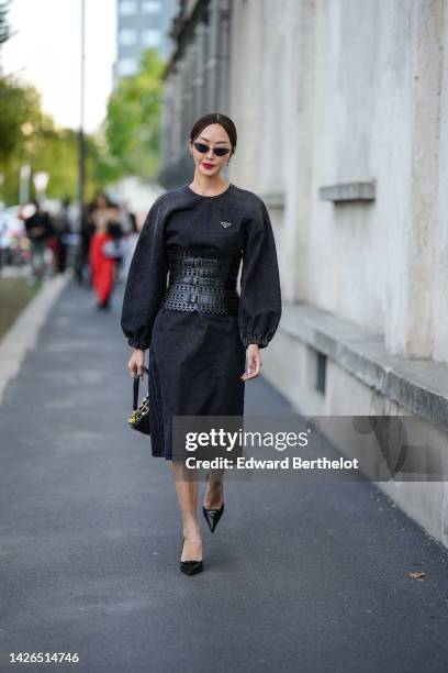 Chriselle Lim wears black cat eyes sunglasses, silver earrings, a black denim long sleeves / knees dress from Prada, a black shiny leather cut-out...