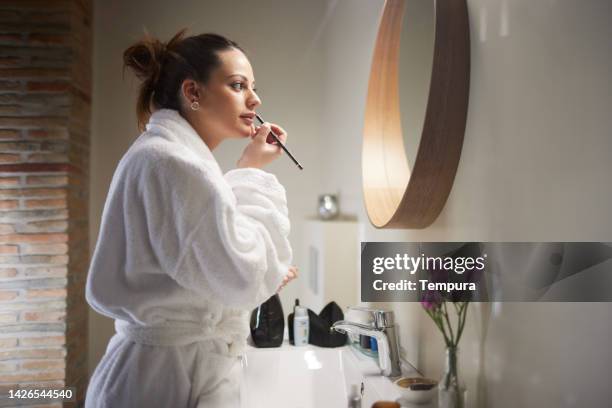 a woman is putting lipstick on. - woman in bathrobe stock pictures, royalty-free photos & images