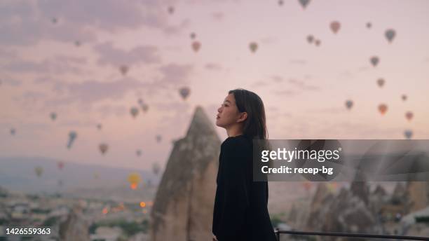 female tourist enjoying watching hot air balloons flying in the sky at rooftop of hotel where she is staying during her vacation - asian woman smiling sunrise stock pictures, royalty-free photos & images