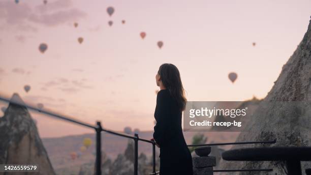 female tourist enjoying watching hot air balloons flying in the sky at rooftop of hotel where she is staying during her vacation - cappadocia hot air balloon stock pictures, royalty-free photos & images