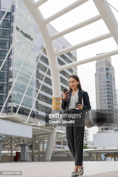 professional smart cool asian young attractive businesswoman holding a cup of coffee while walking and looking at the phone in a business district - bangkok business stock pictures, royalty-free photos & images