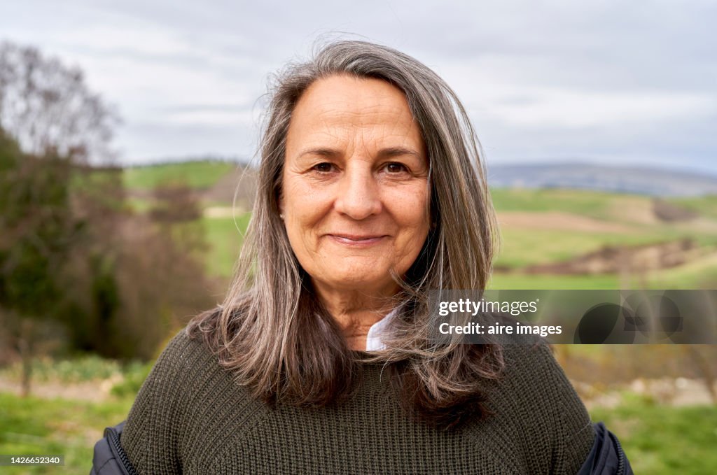 Beautiful older woman with long gray hair enjoying a beautiful autumn day in the countryside, looking at the camera with a smile.