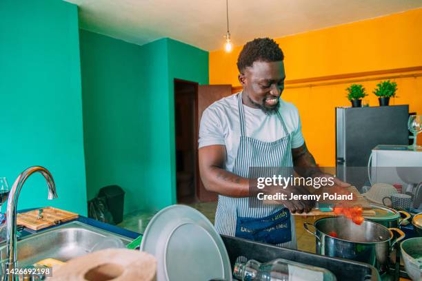 cheerful male chef adding sliced tomatoes to a stew pot in his home kitchen in panama city, panama - kenyan culture stock pictures, royalty-free photos & images