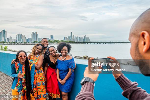 guía turístico local fotografiando a un grupo de turistas afrodescendientes alegres, sonrientes y de moda con una vista del horizonte de la ciudad de panamá por la noche en un día nublado - panamá fotografías e imágenes de stock