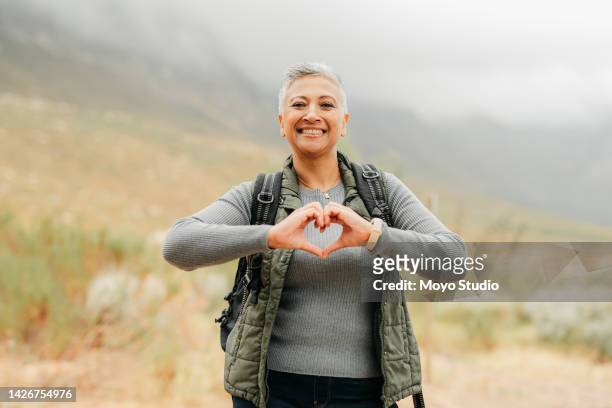 nature hiking senior woman with love and heart sign or hand for healthy, wellness and care on adventure walk on hill, green mountain environment. trekking person with emoji hands, portrait and smile - heart health 個照片及圖片檔