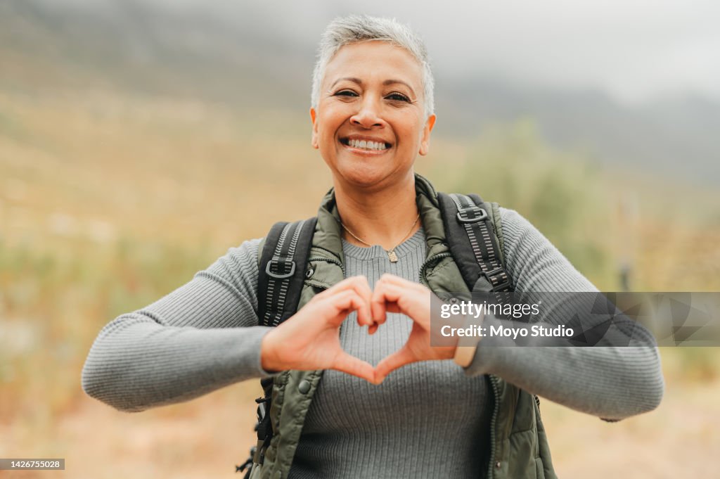 Senior woman with hand heart sign, love for hiking in nature mountains and care for travel adventure on retirement holiday in the countryside. Portrait of elderly person with smile on vacation