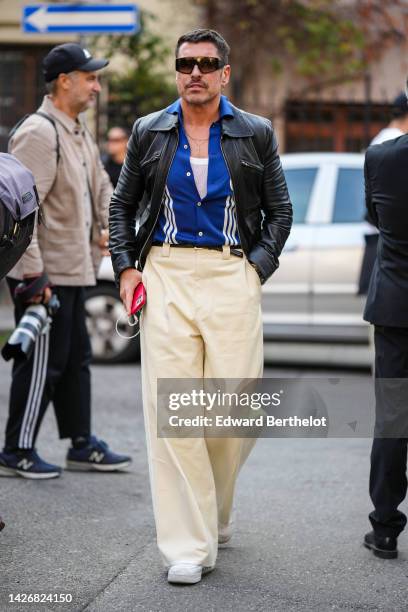 Alex Badia wears black sunglasses from Mom, a white tank-top, a navy blue with white striped print pattern polo shirt, a black shiny leather jacket,...