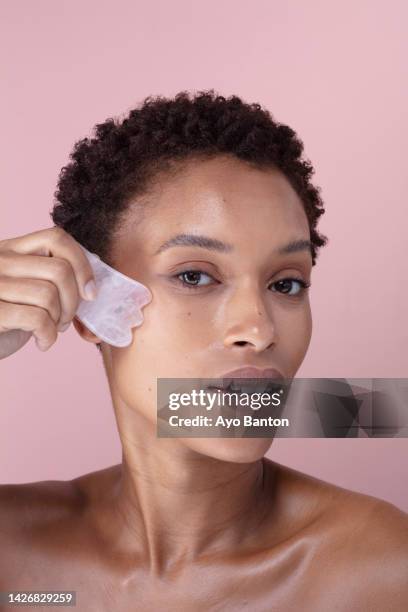 studio portrait of woman massaging face with rose quartz guasha - jadesteine stock-fotos und bilder