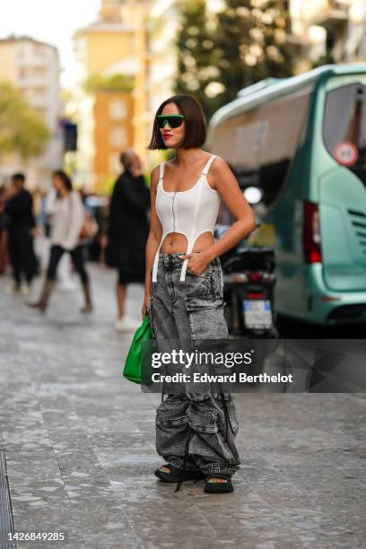 Tiffany Hsu wears a black and neon green futurist sunglasses, a white tank-top / zipper corset with suspender belt, gray faded denim large cargo...