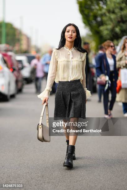 Guest wears a beige tulle ruffled details silk blouse, a beige matte leather shoulder bag, a dark gray tweed pleated short skirt, black shiny leather...